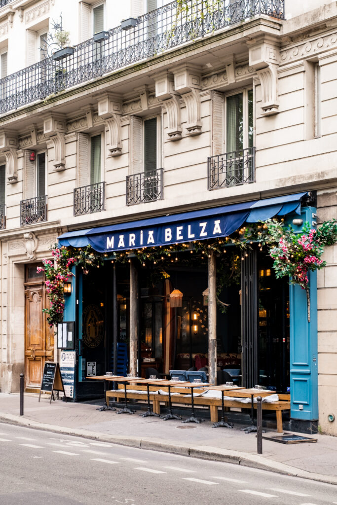 This image shows the exterior of a restaurant named "MARÍA BELZA" with a blue awning and decorative string lights. The entrance is adorned with floral arrangements and a chalkboard menu. There are several tables and benches arranged outside, suggesting outdoor seating. The building has ornate architectural details and balconies with wrought iron railings. A wooden door is visible to the left of the entrance, and there are various signs and decorations around the area.