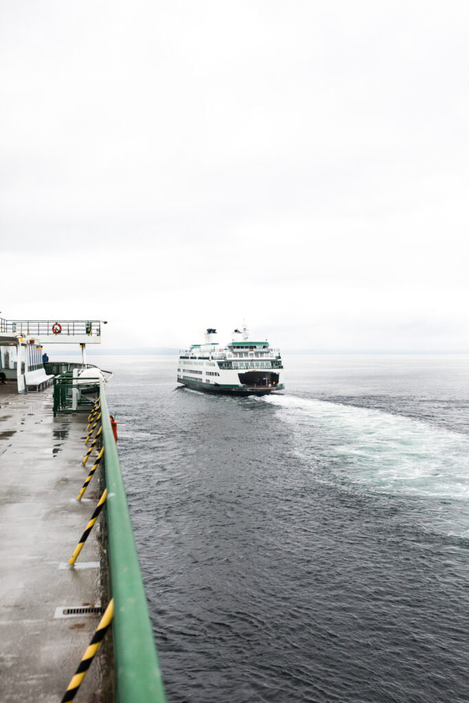 This image shows a ferry boat moving through the water, creating a visible wake behind it. The ferry is white with green accents and has a large open deck area. The scene is captured from a dock, which has a green railing with yellow and black stripes running along its edge. On the dock, there is a lifesaver hanging and a small shelter with benches inside. The water appears calm, and the sky is overcast.