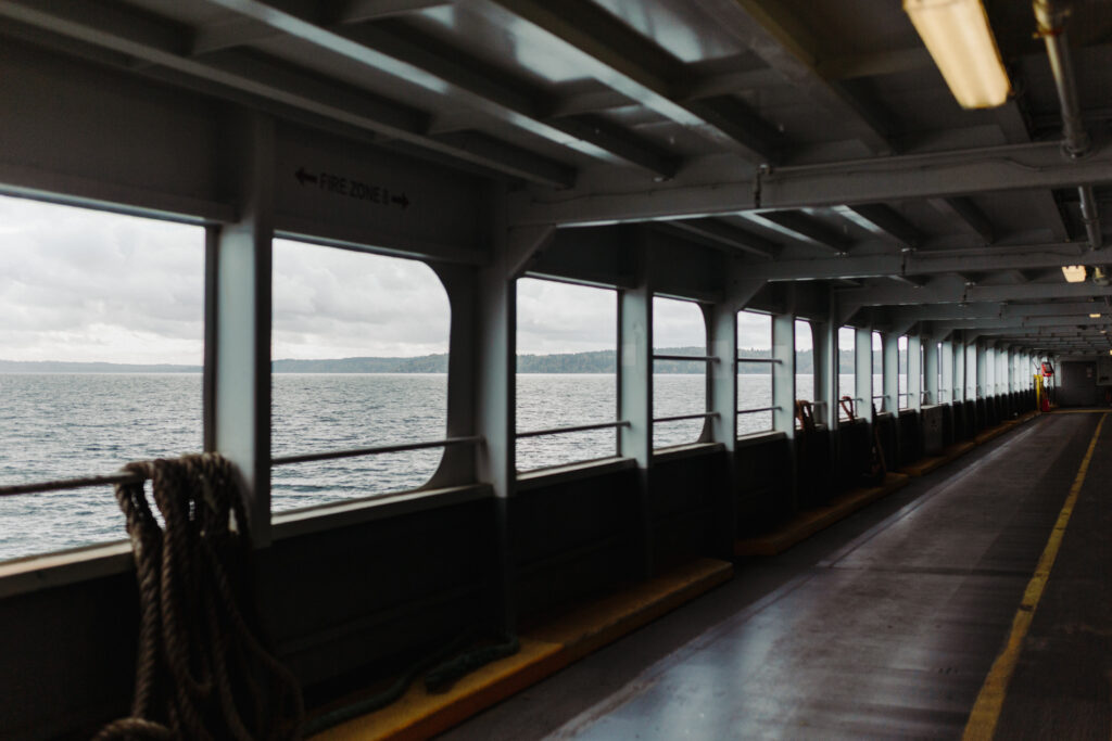 This image shows the interior of a ferry or a large boat with a view of the ocean through multiple rectangular windows. The interior features a long corridor with a yellow line marking the center of the walkway. There are ropes and railings visible near the windows. The ceiling has a grid-like structure with lights and ventilation ducts. A sign indicating "FIRE ZONE" is visible above one of the windows, suggesting safety instructions for passengers. The ocean outside is calm with a horizon line visible in the distance.