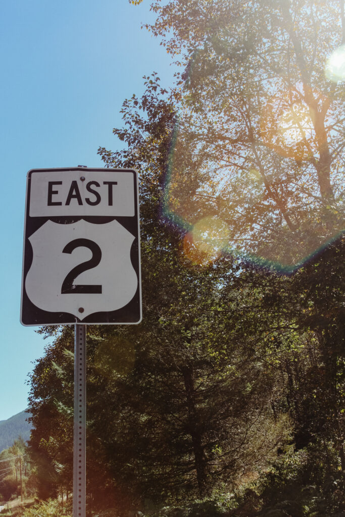 This image shows a road sign indicating "EAST 2" mounted on a metal pole. The sign is positioned against a backdrop of dense trees with sunlight filtering through the foliage, creating a bright and somewhat hazy effect. The sky above is clear and blue. In the distance, there is a glimpse of a mountain range, adding to the scenic view. The road sign is prominently displayed, ensuring it is easily visible to anyone passing by.