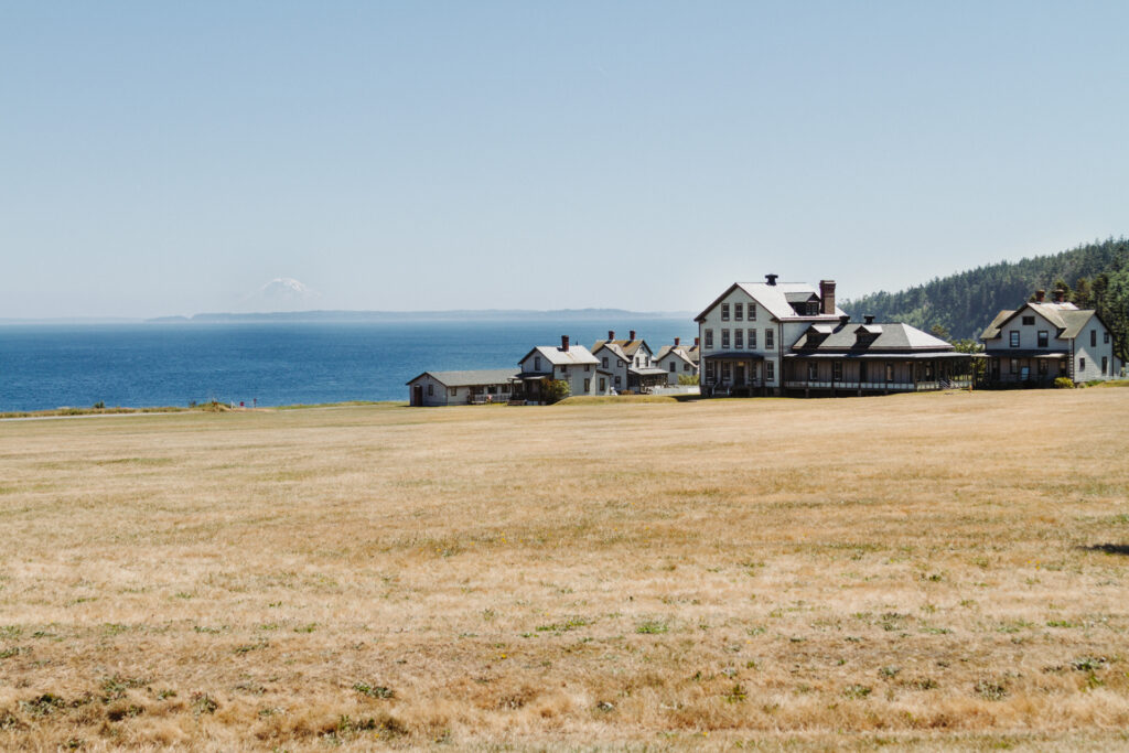 This image shows a serene coastal landscape with a large, open field of dry grass in the foreground. In the background, there are several houses situated near the shoreline, with one prominent larger house standing out among them. The houses are positioned on a slight elevation, providing a picturesque view of the ocean stretching out towards the horizon. The sky is clear and blue, and in the distance, a mountain peak can be seen rising above the horizon line where the ocean meets the sky. The overall scene conveys a tranquil and idyllic setting.