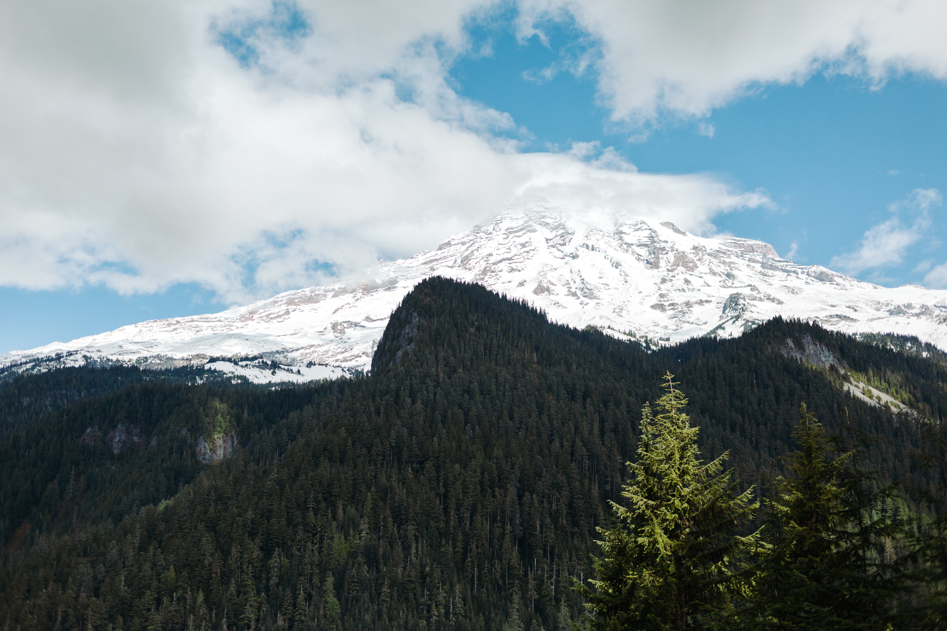 This image shows a breathtaking view of a snow-capped mountain peak under a partly cloudy sky. The mountain is covered in snow, with patches of green forest at its base. The forest is dense with tall trees, and the foliage appears lush and vibrant. The sky above is a mix of blue and white clouds, suggesting fair weather. The overall scene is serene and picturesque, capturing the natural beauty of a mountainous landscape.
