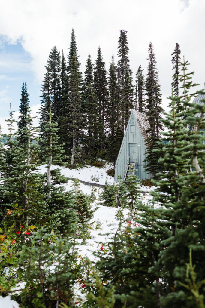 This image shows a serene and picturesque scene of a small, triangular-shaped cabin nestled among tall evergreen trees in a snowy forest. The cabin has a slanted roof and a wooden door, with a ladder leaning against it. The surrounding area is covered with snow, and there are various green coniferous trees and shrubs dotting the landscape. The sky above is partly cloudy, with patches of blue visible. The overall atmosphere is tranquil and inviting, suggesting a remote and peaceful location.