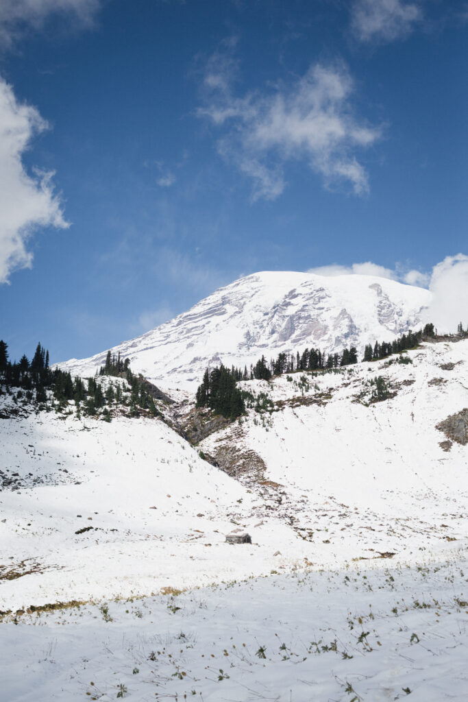 This image shows a serene and picturesque winter landscape dominated by a snow-covered mountain peak under a clear blue sky with scattered clouds. The foreground features a snow-covered slope with patches of grass peeking through the snow. The middle ground includes a series of snow-covered hills and ridges, with evergreen trees dotting the landscape. The mountain peak, likely a well-known landmark given its prominence, is completely blanketed in snow, suggesting a cold and possibly harsh environment. The overall scene conveys a sense of tranquility and natural beauty.