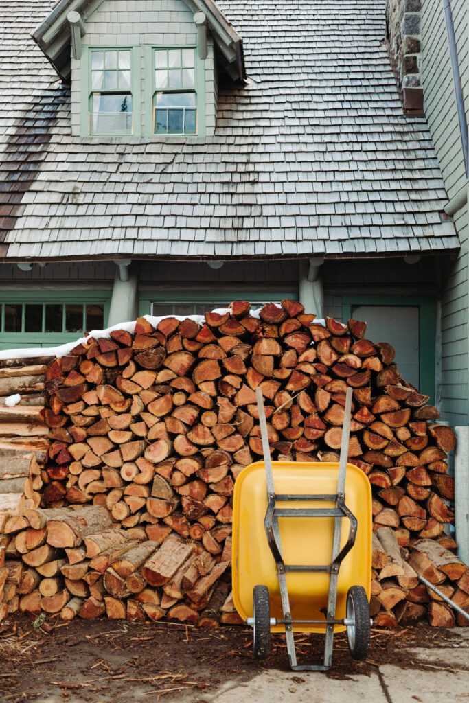 This image shows a large pile of firewood stacked neatly in front of a house. The firewood consists of numerous logs arranged in an orderly fashion. In front of the pile, there is a yellow wheelbarrow with a metal frame and two wooden handles. The house has a gray shingle roof and green-framed windows. The scene appears to be set in a rural or suburban area, with the house and the firewood suggesting preparation for colder weather.