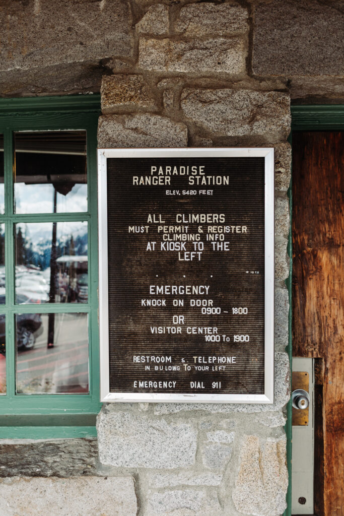This image shows a sign attached to a stone wall, providing information for visitors at the Paradise Ranger Station. The sign includes details such as the elevation of the station, instructions for climbers to permit and register at the kiosk to the left, emergency contact information, and directions to the visitor center and restrooms. The sign is framed in white and mounted on the textured stone wall, with a green-framed window to the left reflecting the outdoor scene, which includes parked cars and a distant view of trees and possibly mountains.