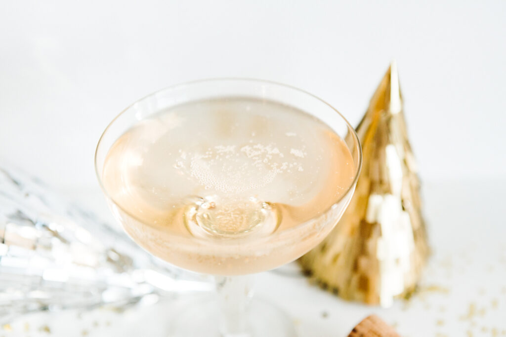 This image shows a glass of champagne with a golden cone-shaped decoration to the right of it. The champagne is clear with visible bubbles and a slight golden hue. The glass is positioned on a white surface, and there is a cork from the champagne bottle lying nearby. The overall setting suggests a celebratory atmosphere.