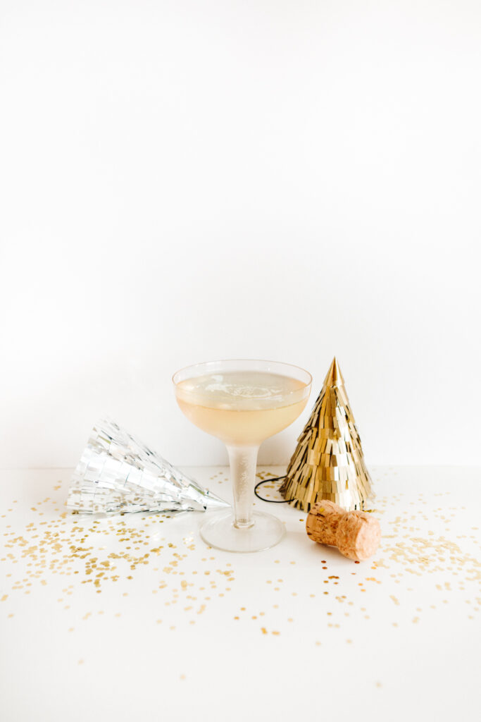 This image shows a clear cocktail glass filled with a light-colored beverage, possibly champagne or a similar sparkling drink. Next to the glass, there is a gold-colored party hat and a silver party horn, suggesting a celebratory occasion. A cork from a champagne bottle is also present on the table. The table is covered with a white tablecloth, and there are gold confetti scattered around, enhancing the festive atmosphere.