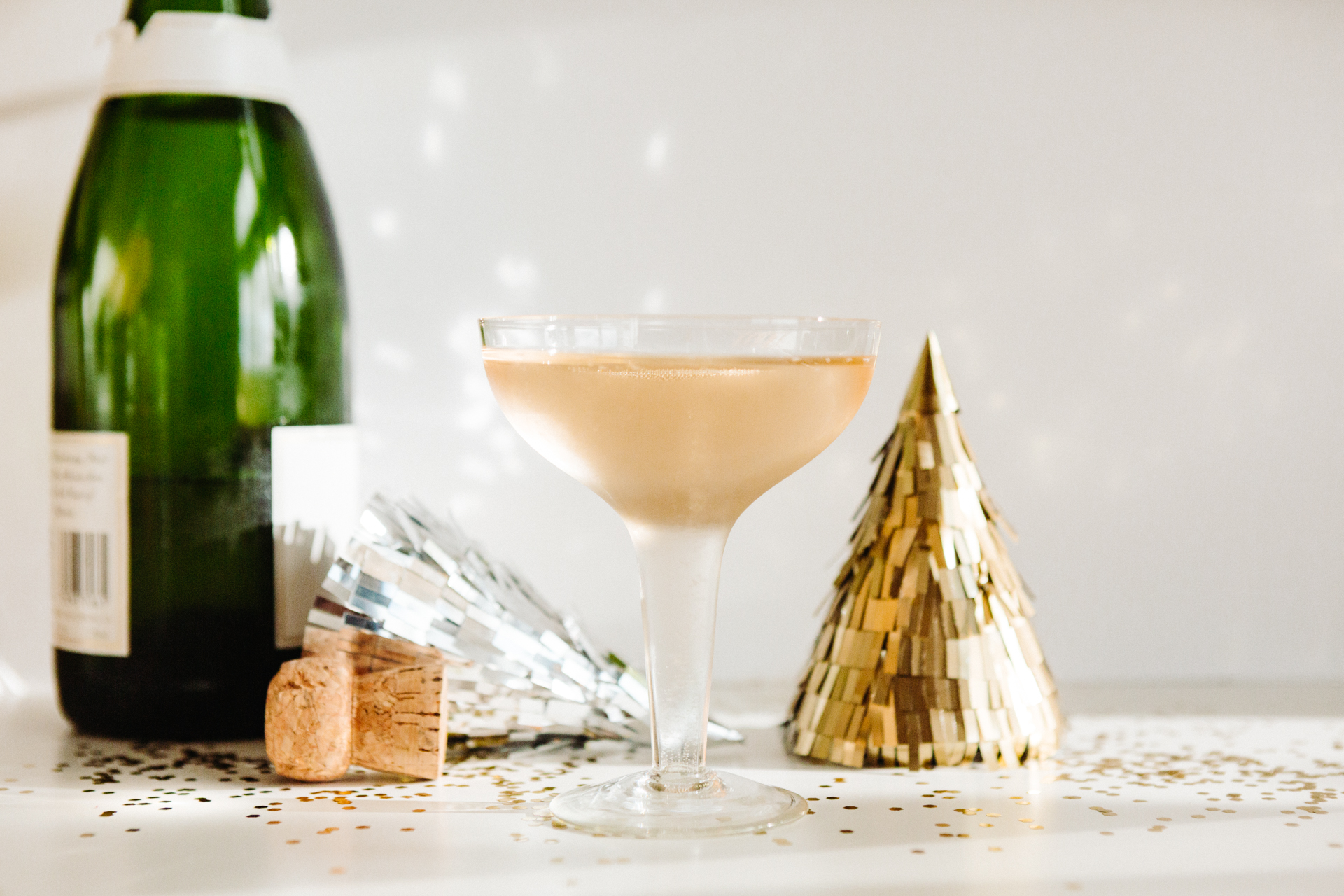 This image shows a festive scene with a glass of champagne, a green champagne bottle, and celebratory decorations. The champagne glass is filled with a light-colored liquid and is placed on a white surface. Next to the glass, there is a green champagne bottle with a white label. To the right of the glass, there is a gold foil party hat. Additionally, there are silver party decorations and a cork on the table. The table is covered with gold confetti, adding to the celebratory atmosphere.