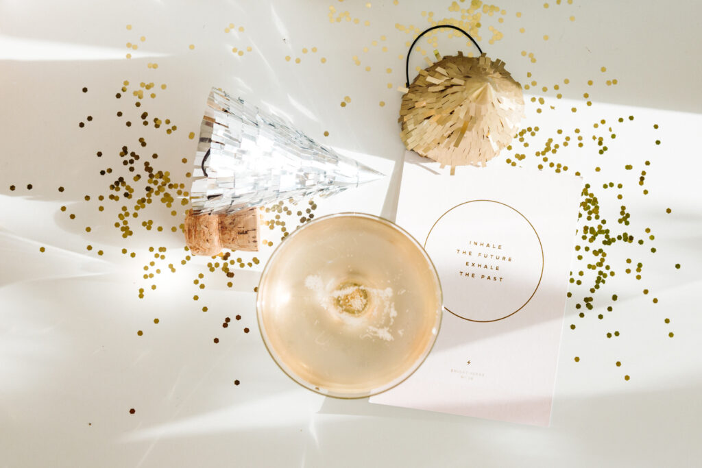 This image shows a festive scene with various celebratory elements. There is a champagne glass filled with a light-colored beverage, placed next to a cork from a champagne bottle. A metallic silver party hat is positioned behind the glass. To the right, there is a decorative gold piñata hanging from a black string. Scattered gold confetti dots the white surface, adding to the celebratory atmosphere. A piece of paper with printed text is partially visible, featuring phrases like "Inhale the Future" and "Exhale the Past," suggesting a theme of moving forward or celebrating new beginnings.